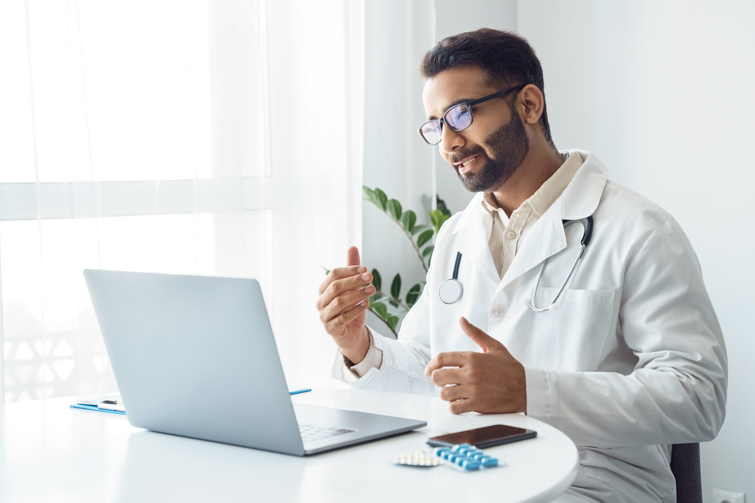 Portrait of indian man doctor talking to online patient on laptop screen sitting at clinic office desk giving online consultation for domestic health treatment. Telemedicine remote medical appointment